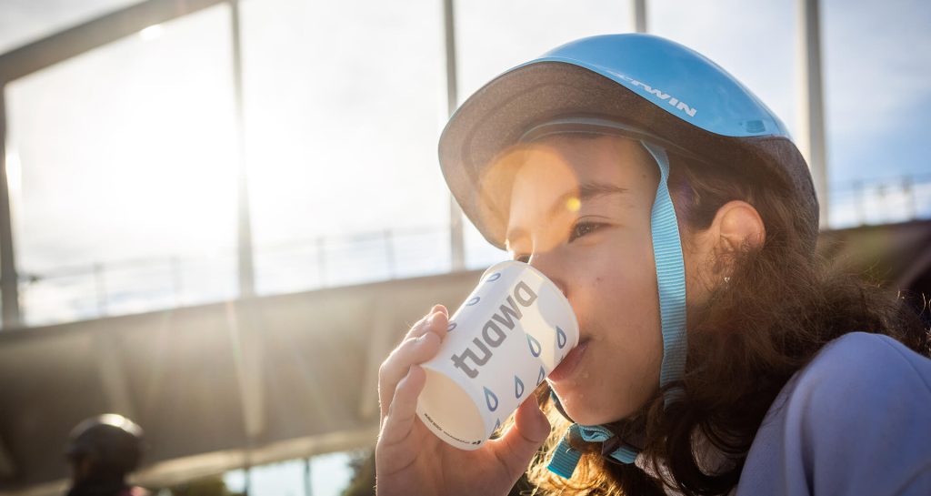 Niña con casco de bicicleta bebiendo agua sostenible en vaso Tuawa en evento al aire libre