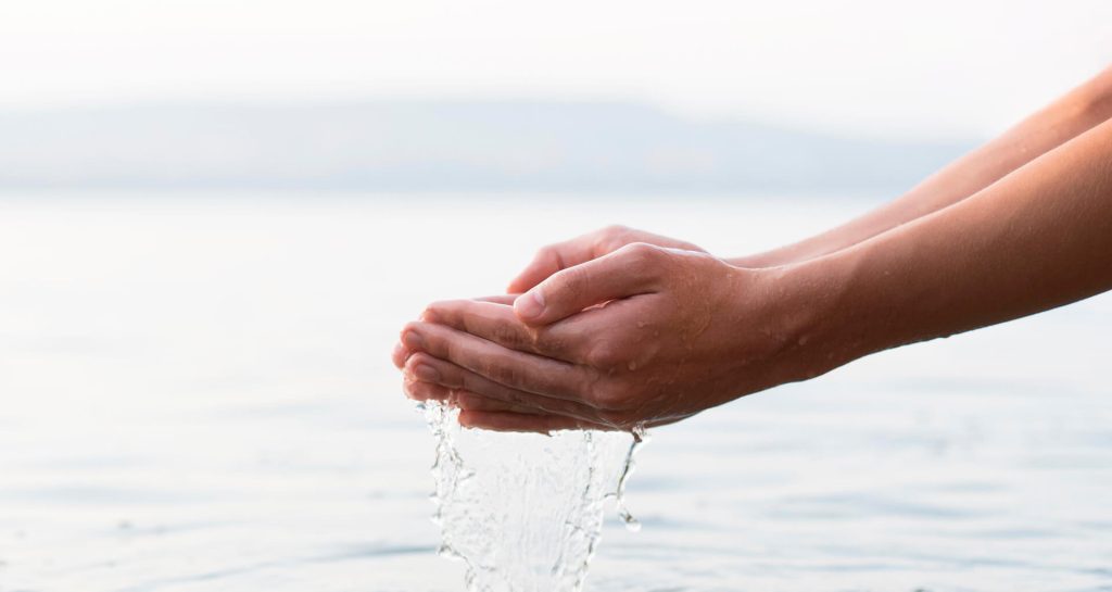 Manos recogiendo agua cristalina con el mar al fondo, símbolo de hidratación y sostenibilidad Tuawa